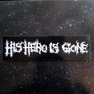 Op de afbeelding: Zwart-witte sticker met de tekst "HIS HERO IS GONE" in een gestileerd lettertype.