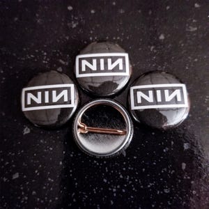 May include: Three circular pin-back buttons featuring a black background with a white rectangular border containing the text NIN in bold white capital letters. One button is flipped over to reveal a metallic pin mechanism on the back.