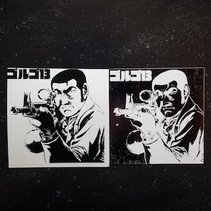 Op de afbeelding: Twee vierkante stickers met een zwart-wit illustratie van een man die richt met een geweer met vizier. De Japanse tekst "ゴルゴ13" staat bovenaan elke sticker. Eén sticker heeft een witte achtergrond, de andere een zwarte achtergrond.