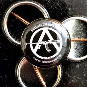May include: Black and white button with an anarchy symbol and a rifle inside. The text "MASSLESS CREATION" and "MALICIOUS CORROSIVE INSURRECTION" is around the symbol. The text "M.C.I." is in the center of the button.