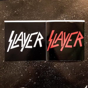 May include: Two black and white stickers with the word "SLAYER" in a stylized font. The sticker on the left has white lettering and the sticker on the right has red lettering.