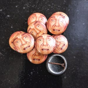 May include: A set of eight round button pins featuring a close-up image of a man's face. The buttons are arranged in a pile on a black surface.