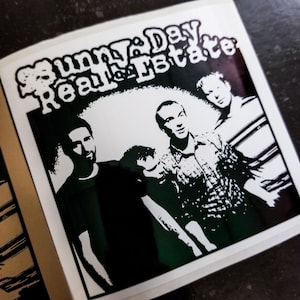 May include: Black and white photo of a band called "Sunny Day Real Estate". The band members are standing in front of a white background. The band's name is written in a bold, stylized font.