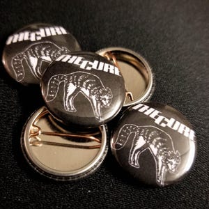 May include: Four black and white button badges featuring the word "The Cure" and a stylised image of a cat. The badges are arranged in a pile.