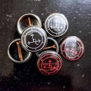 May include: A group of six black and white button badges with a white sigil on a black background. One badge has a red border around the sigil.