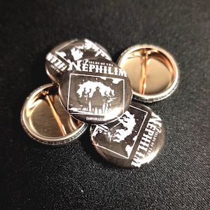 May include: Four black and white button pins with the text "Fields of the Nephilim" and the band's logo. The pins are on a black surface.