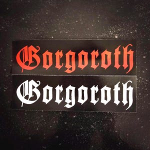 Może przedstawiać: Dwa czarno-białe naklejki z napisem "Gorgoroth" napisanym czcionką gotycką. Górna naklejka ma napis w kolorze czerwonym, a dolna naklejka ma napis w kolorze białym.