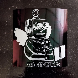 May include: Black and white sticker featuring a robot with angel wings wearing headphones. The robot is sitting with its legs crossed and has a large, round head with two large eyes. The text "THE GET UP KIDS" is printed below the robot.