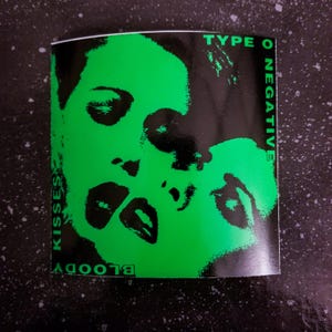 May include: A black and green sticker with the text "TYPE O NEGATIVE" and "BLOODY KISSES" in white. The sticker features a black and white image of a woman's face.