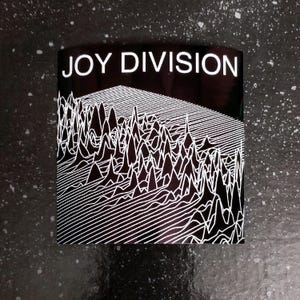 Op de afbeelding: Zwart-wit grafisch ontwerp met de tekst "JOY DIVISION" bovenaan. Het ontwerp bevat een reeks witte lijnen die een golvend patroon creëren.