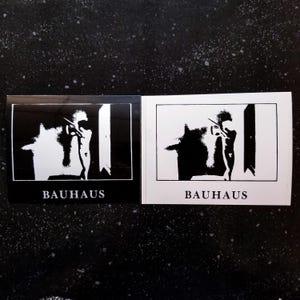 Könnte beinhalten: Zwei schwarz-weiße Aufkleber mit dem Wort "BAUHAUS" in weiß auf schwarzem Hintergrund. Jeder Aufkleber zeigt die Silhouette einer Person, die in einer Tür steht.