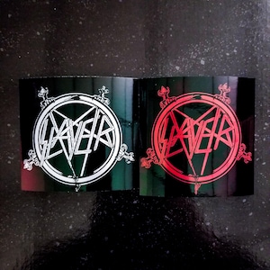 May include: Two black square stickers with the Slayer logo. The logo is a circle with a pentagram inside and the word "Slayer" written in a stylized font. One sticker has a white logo and the other has a red logo.