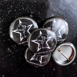 Peut inclure: Plusieurs badges noirs et blancs avec un profil stylisé dans une étoile et le texte "THE SISTERS OF MERCY". L'un des badges montre l'arrière de l'épingle. Les badges sont disposés sur une surface sombre et texturée.