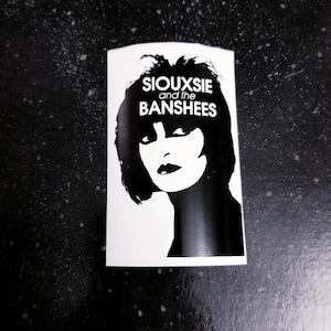 May include: Black and white sticker featuring a portrait of Siouxsie Sioux, lead singer of the band Siouxsie and the Banshees. The text "Siouxsie and the Banshees" is printed in white letters on a black background.