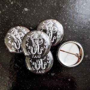 Puede incluir: Cuatro pines de botón negros y blancos con un gráfico de una criatura con cola de serpiente y el texto "IAW CEWC" alrededor del borde. Un pin de botón negro liso con un pasador de metal.