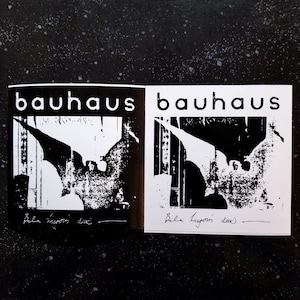 May include: Two black and white stickers with the band name "Bauhaus" printed in white letters. The stickers feature a black and white image of a bat with a person's face in the center. The text "Bela Lugosi Dead" is written in black ink at the bottom of each sticker.
