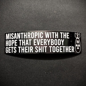 Op de afbeelding: Zwarte rechthoekige sticker met witte tekst: "MISANTHROPIC WITH THE HOPE THAT EVERYBODY GETS THEIR SHIT TOGETHER." Een kleine afbeelding van een bom met een stralingssymbool.