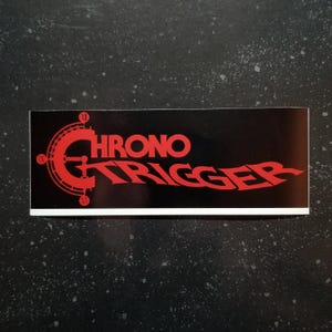 Peut inclure: Un autocollant noir et rouge avec les mots "Chrono Trigger" dans une police stylisée. Le logo présente un cadran d'horloge avec des chiffres romains.