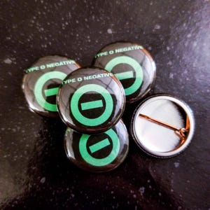 以下が含まれることがあります： 「Type O Negative」という文字と、黒い水平線が入った緑色の円が描かれた、黒と緑の丸いボタンが5つ。ボタンの1つは、銀色のピンが付いた裏側です。