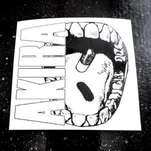 Op de afbeelding: Een zwart-witte sticker met het woord "AKIRA" in vette, omrande letters. De afbeelding toont een open mond met tanden en twee pillen erin. De sticker staat op een witte achtergrond.