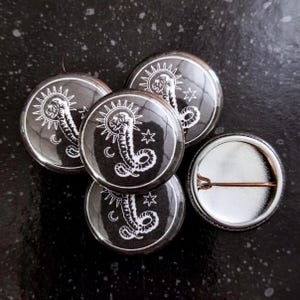 May include: Four black and white button badges featuring a snake design with a sun and moon behind it. The fourth badge is a blank back.