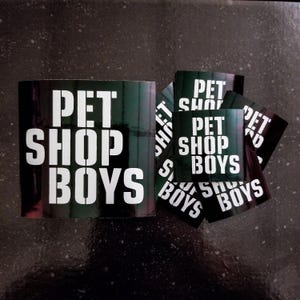 May include: A set of black and green stickers with the text "PET SHOP BOYS" in white letters. The stickers are arranged in a stack, with the largest sticker on the bottom and the smallest sticker on top.