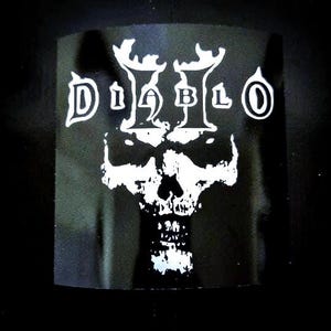 Puede incluir: Diseño gráfico en blanco y negro de una calavera con el texto "DIABLO II" encima.