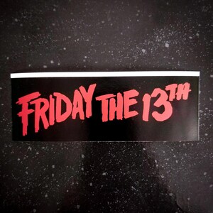 Può includere: Un adesivo nero e bianco con il testo rosso "FRIDAY THE 13TH" in un font stilizzato.