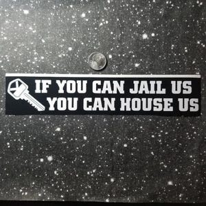 Könnte beinhalten: Schwarzer Aufkleber mit weißem Text: "IF YOU CAN JAIL US YOU CAN HOUSE US". Eine weiße Schlüsselgrafik befindet sich links. Eine Münze befindet sich darüber.