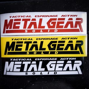 May include: Three stickers with the text "Tactical Espionage Action Metal Gear Solid" in different color combinations. The first sticker is red on white, the second is black on yellow, and the third is white on black.