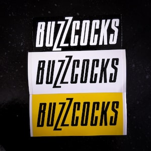 May include: Three stickers with the word "Buzzcocks" in black letters on a white background, a white background, and a yellow background.