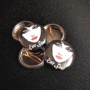 May include: Four black and white button pins with a portrait of Kate Bush and the text "Kate Bush" in white lettering.