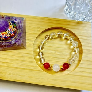 May include: A clear and gold beaded bracelet with red and white accent beads. The bracelet is displayed on a wooden tray with a clear circular indentation. A purple and gold decorative object is in the background.