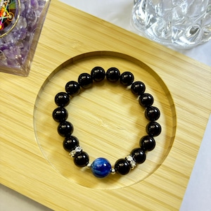 Pode incluir: Pulseira de contas pretas com uma única conta azul e prateada. A pulseira é feita de contas pretas redondas e polidas e apresenta uma conta azul central. A pulseira é exibida em uma bandeja de madeira.