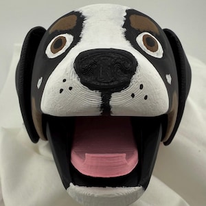 May include: A 3D printed dog head puppet with a black, white, and brown painted design. The puppet has a large, open mouth with a pink tongue.