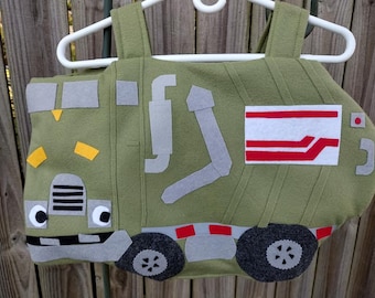 May include: An olive green felt costume shaped like a garbage truck, featuring gray felt wheels, a front grille with white eyes, yellow accents, and a rectangular white patch with red stripes on the side, all suspended from a white plastic hanger.