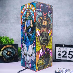 Puede incluir: Lámpara rectangular con ilustraciones estilo vidriera que representan personajes de fantasía. La lámpara tiene un cable negro y está decorada con imágenes de una mujer, un demonio, un lobo y un dragón. La lámpara está encendida.