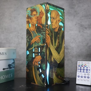 Peut inclure: Lampe rectangulaire avec une illustration de deux personnages et un chat, dans des tons de vert, or et bleu. La lampe émet une lueur bleue. Une télécommande est également visible.