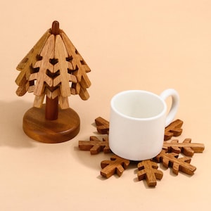 May include: A set of two wooden coasters, one shaped like a Christmas tree and the other shaped like a snowflake. The coasters are made of natural wood and have a rustic look. The tree coaster is folded and has a small base. The snowflake coaster is flat and has a six-pointed star shape.