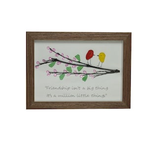 May include: Framed artwork featuring two glass birds perched on a branch with pink and green glass flowers. The quote "Friendship isn't a big thing. It's a million little things." is written below the birds.