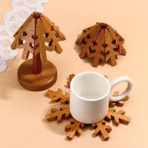 Könnte beinhalten: Holzuntersetzer in Baumform und eine weiße Keramiktasse. Die Untersetzer sind aus hellbraunem Holz gefertigt und als stilisierte Bäume gestaltet. Ein Untersetzer steht auf einem Ständer, ein weiterer ist flach, der dritte unter der Tasse.
