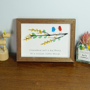 May include: A framed artwork with a quote about friendship. The artwork features a branch with yellow and green glass beads, and two birds made of glass beads. The quote reads: "Friendship isn't a big thing. It's a million little things."