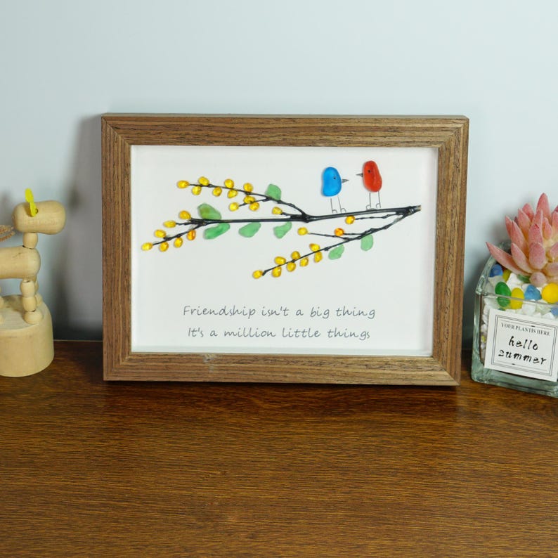 May include: A framed artwork with a quote about friendship. The artwork features a branch with yellow and green glass beads, and two birds made of glass beads. The quote reads: "Friendship isn't a big thing. It's a million little things."