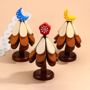 May include: Three wooden Christmas tree decorations with different coloured tops. The first tree has a blue crescent moon with white stars, the second tree has a red flower with a white snowflake, and the third tree has a yellow crescent moon with white stars.