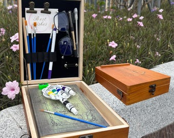 May include: A wooden art box, open to reveal art supplies. The box contains paint tubes, brushes, a palette, and a notebook. The lid has a black interior with pens, glasses, and a card that says "Best wishes".