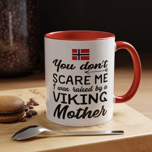Peut inclure: Un mug en céramique blanche avec un intérieur et une anse rouges, arborant le drapeau norvégien et le texte noir : You don't SCARE ME I was raised by a VIKING Mother. Le mug est posé sur une surface en bois à côté d'une petite cuillère et d'un biscuit.