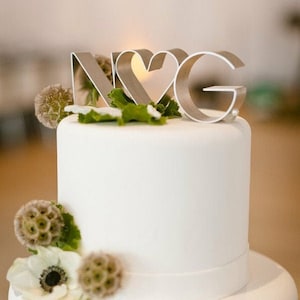 May include: A two-tiered white wedding cake decorated with white flowers and greenery. The top tier has a metal topper with the letters "N" and "G" with a heart in the middle.