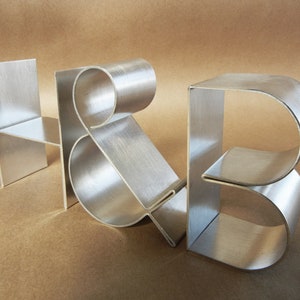 May include: Silver metal sculptures spelling out "H & B" against a brown backdrop. The "H" is a simple, angular design, while the "&" and "B" have a curved, flowing shape. These sculptures are likely decorative items for a home or office.