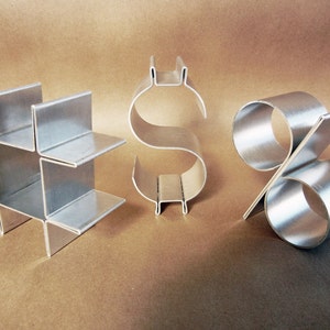 Puede incluir: Tres esculturas metálicas de los símbolos #, $ y %. Las esculturas están hechas de metal de color plateado y están dispuestas en fila sobre una superficie marrón.