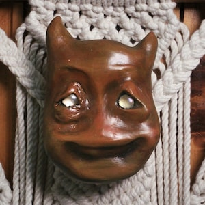 Könnte beinhalten: Eine braune Teufelsmaske mit Hörnern und einem lächelnden Gesicht, mit edelsteinartigen Augen. Sie ist an einem Makramee-Wandbehang aus dickem, cremefarbenem Seil befestigt, der an einem Holzstab hängt. Die Maske hat ein einzigartiges, handgefertigtes Aussehen.